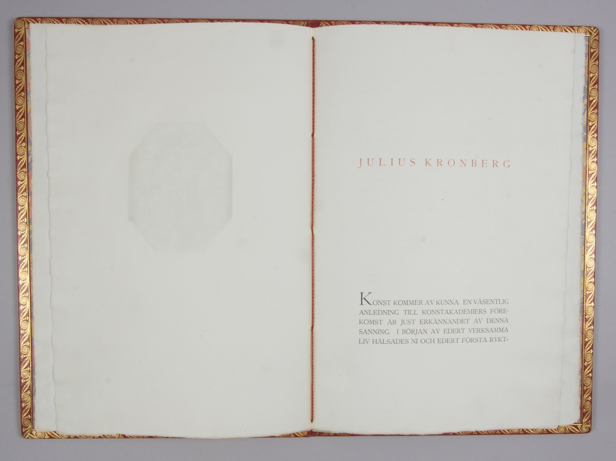 Adress, hyllningsadress, rektangulär i rött skinnband med Konstakademiens emblem präglat i guld på framsidan. Invändigt hyllningstext till Julius Kronberg undertecknad av I. G. Clason, samt kommentar till texten av Julius Kronberg.