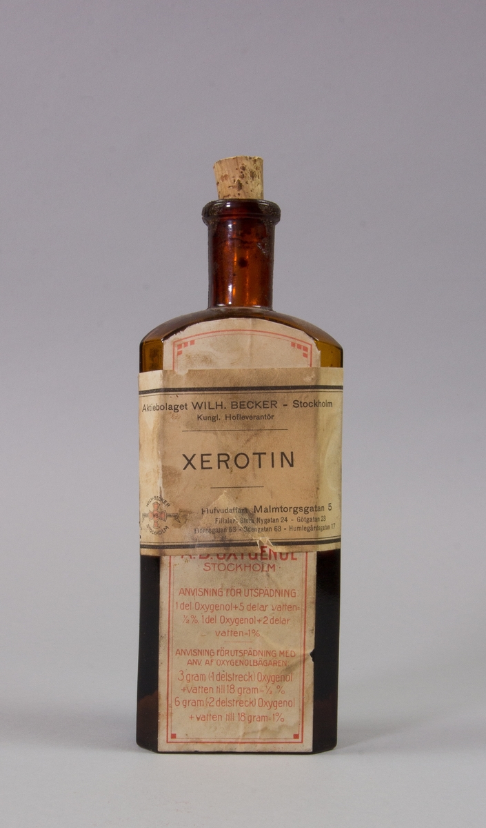 Flaska av brunt glas, rektangulär med kork för xerotin. Har ursprungligen innehållit oxygenol. Fylld tll hälften med xerotin.
