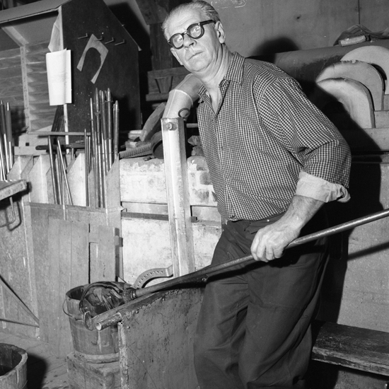 Foto av en man med glasögon som sitter på en glasarbetarstol och ena hand med en blåsrör, arbetar och sysslar med att forma glas.