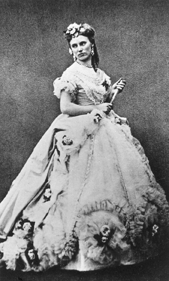 Rollporträtt (?) av Christina Nilsson. Hon bär en stor klänning samt håller i en hopslagen solfjäder. Hon har även blommor i håret. Violetta i La Traviata.