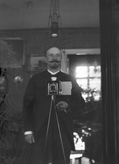 Foto av en skägg- och mustaschprydd herre med kamera, uppställd i ett vardagsrum. 
(Trol. Nils Danckwardt själv).