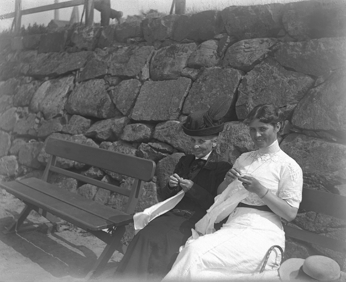 Foto av två kvinnor i olika ålder, som sitter och handarbetar på en parksoffa, troligen vid strandpromenaden i Varberrg.
