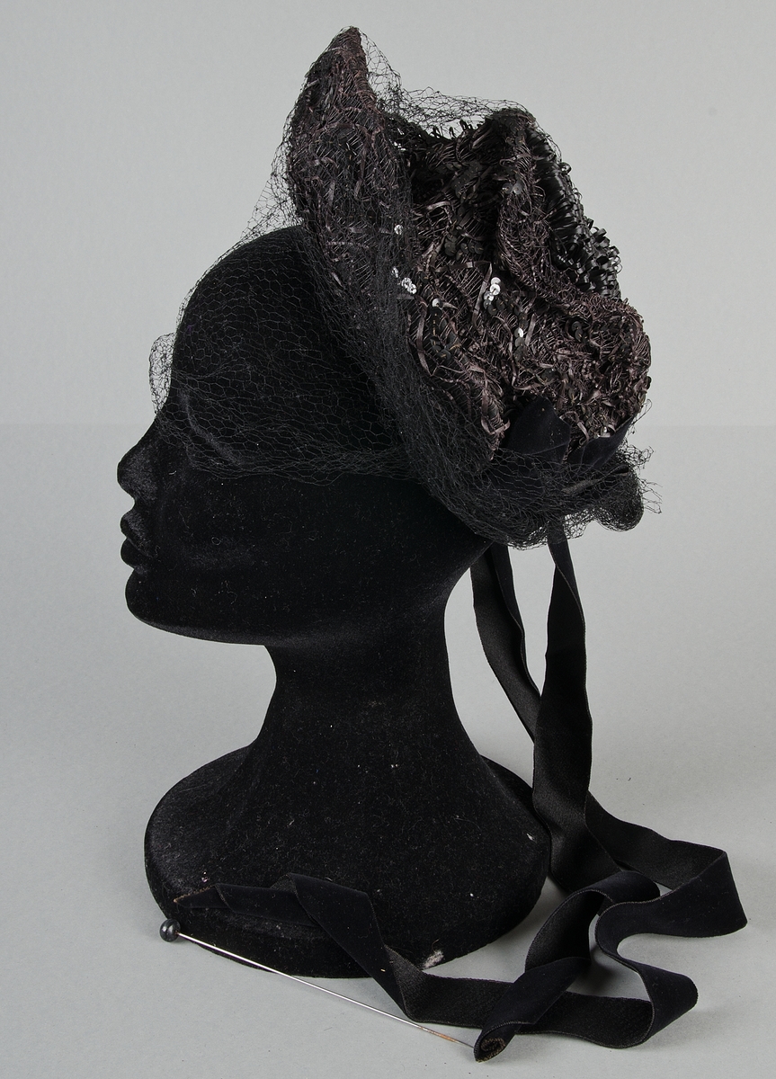 Damhatt av babymodell av svart strå med svart flor, påsydda svarta paljetter, svarta sammetsrosetter och band. Knytband av svart sammet. Fodrad med svart siden.