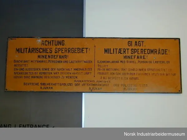 Rektangulært skilt med regler for sort skrift om ferdsel i militært område og advarsel om minefare. Tekst er skrevet på tysk og norsk.