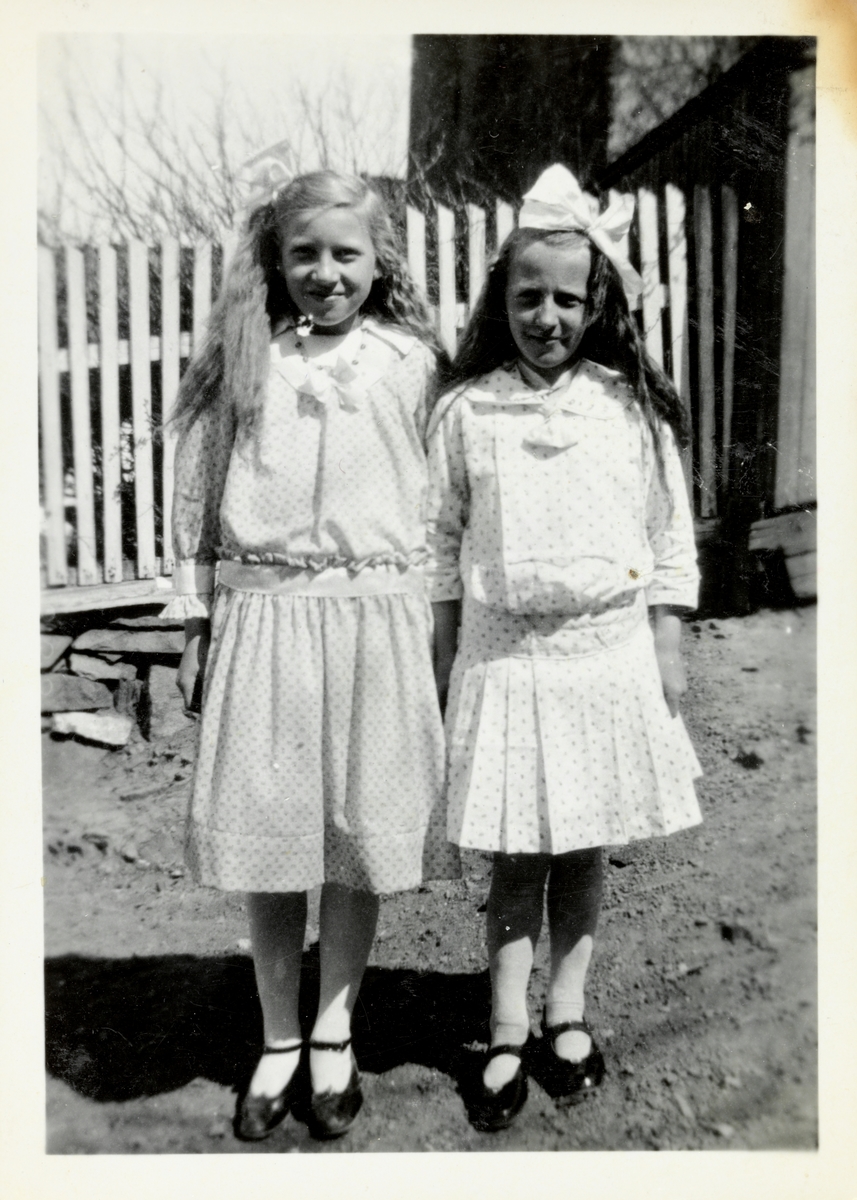 To jenter ikledd kjole poserer foran et stakittgjerde.