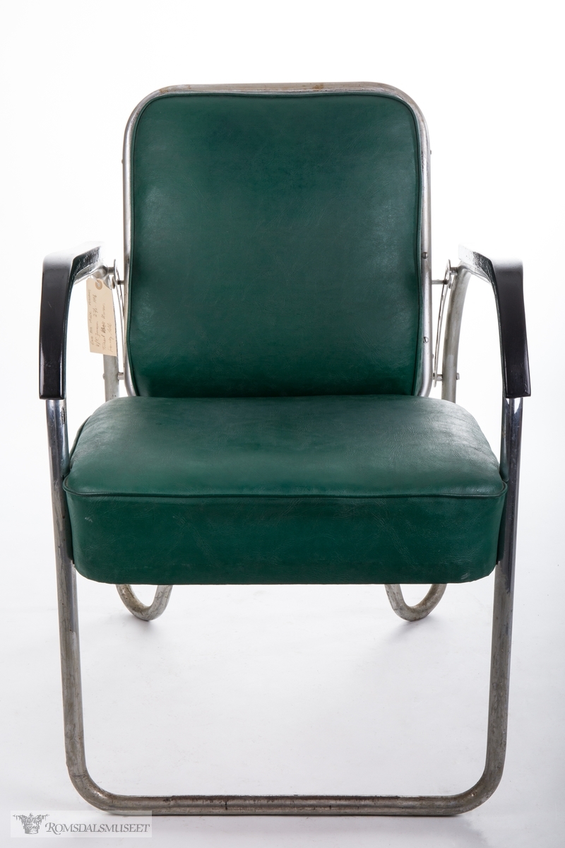 Frisørstol med ramme og ben av metallrør, med reguleringsmekanisme i ryggen. Påsatte armlener i sortlakkert tre. Sete og ryggeputene er trukket i grønt kunstskinn.