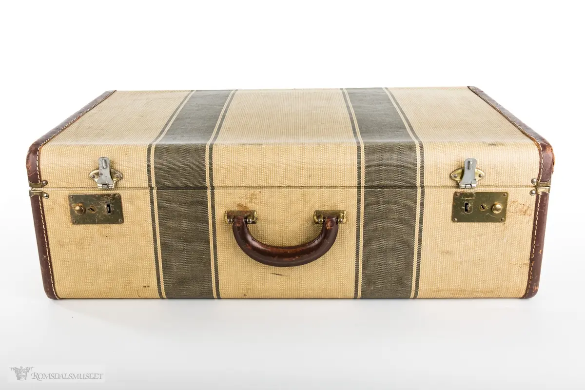 Rektangulær koffert i herdet lerret med forsterkninger av skinn, skinnhåndtak og beslag og lås i messing. Kofferten er kleed med lilla nylonstoff og fire rysjede lommer er sydd inn i trekket på innsiden av lokket og på koffertens sider.