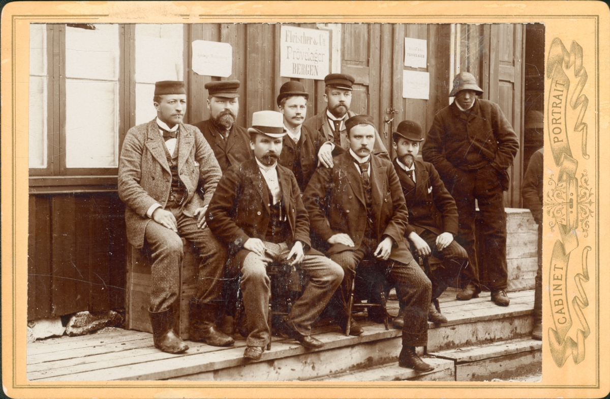 Gruppe.Åtte personer sitter utenfor et lager, sannsynligvis i Bergen. Hans A. Meyer i bakerste rekke med skalk. Det står Fleischer & Co - Prøvelager på et skilt på veggen. Antagelig ca 1880-1890.