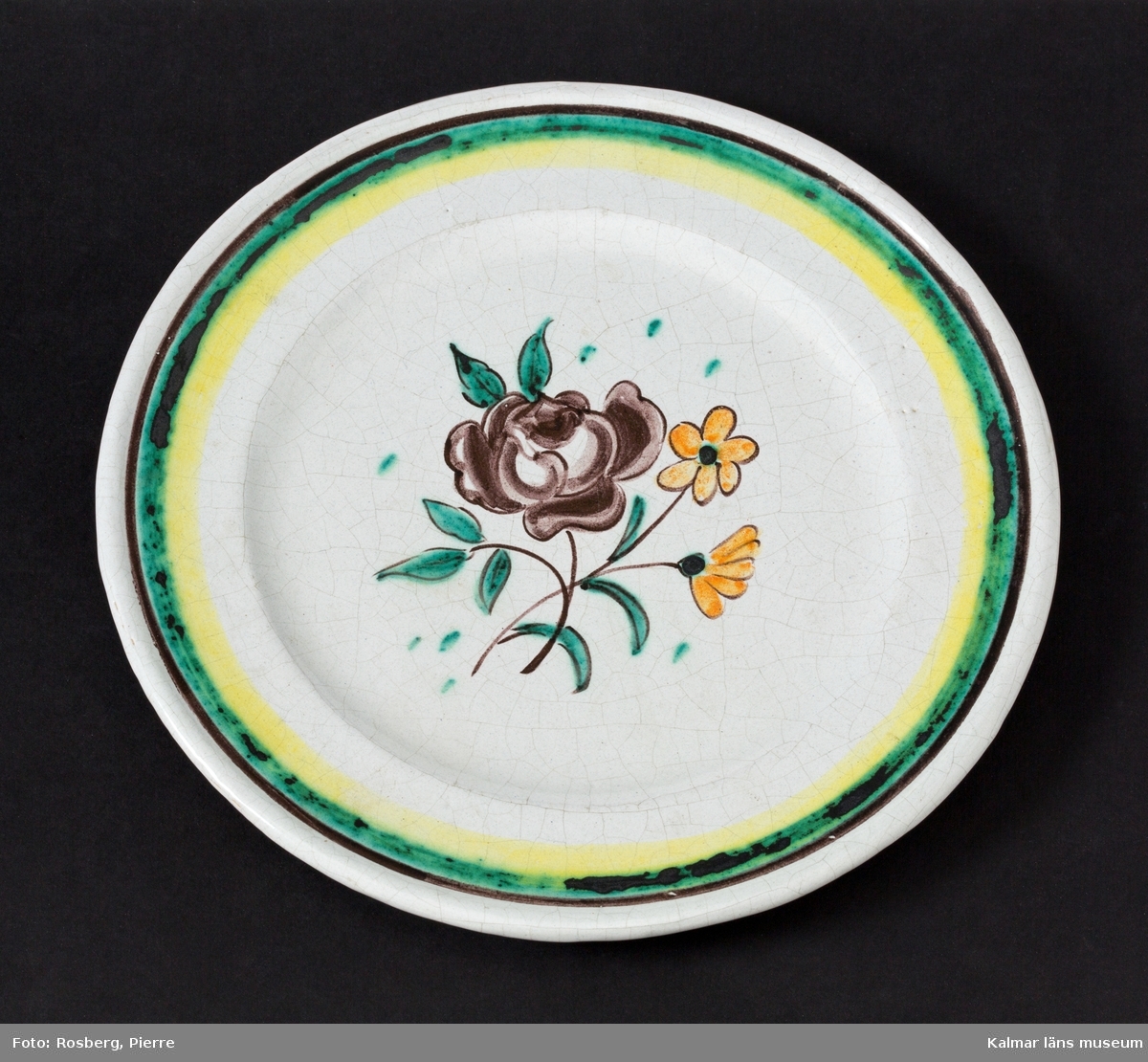 KLM 45844:2 Fat, av keramik. Runt, med blomma i lila, rosa och grönt, grön rand på brättet. Signerad, J.S. Målad och signerad av John Sjöstrand, tillverkad vid Sandbäcks kakelfabrik AB. Dateras till slutet på 1920-talet till 1931.