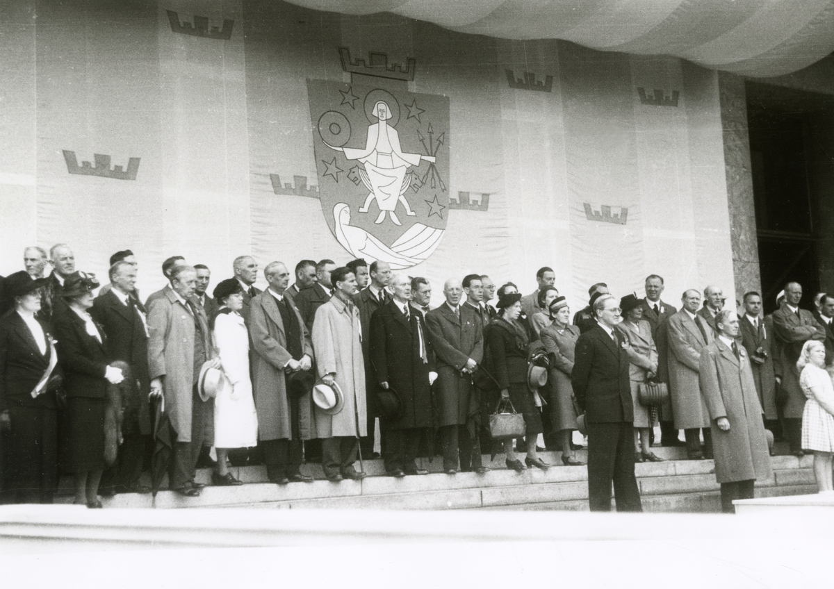 Fra Oslo 7. juni 1945.
Kongen kommer tilbake. Fra velkomsten utenfor Rådhuset.
De prominente gjestene med Oslo Byvåpen i bakgrunnen.