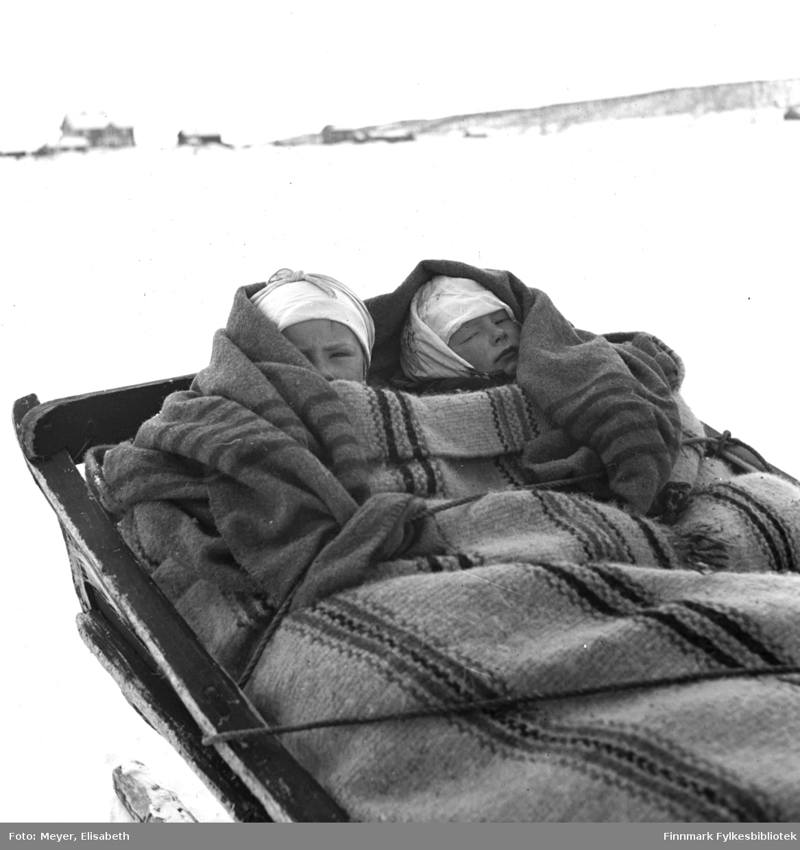 På Sledetur. To barn godt pakket inn i tepper ligger i slede. Det ene teppet er håndvevd i et karakteristsk stripemønster. Fotografert ved påsketider 1940.
