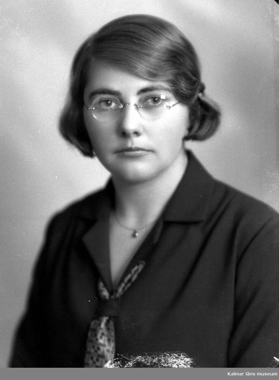 Ateljébild på en kvinna i klänning, halsband, slips och glasögon. Beställare till bilden: Elise Thernström ifrån Kalmar.