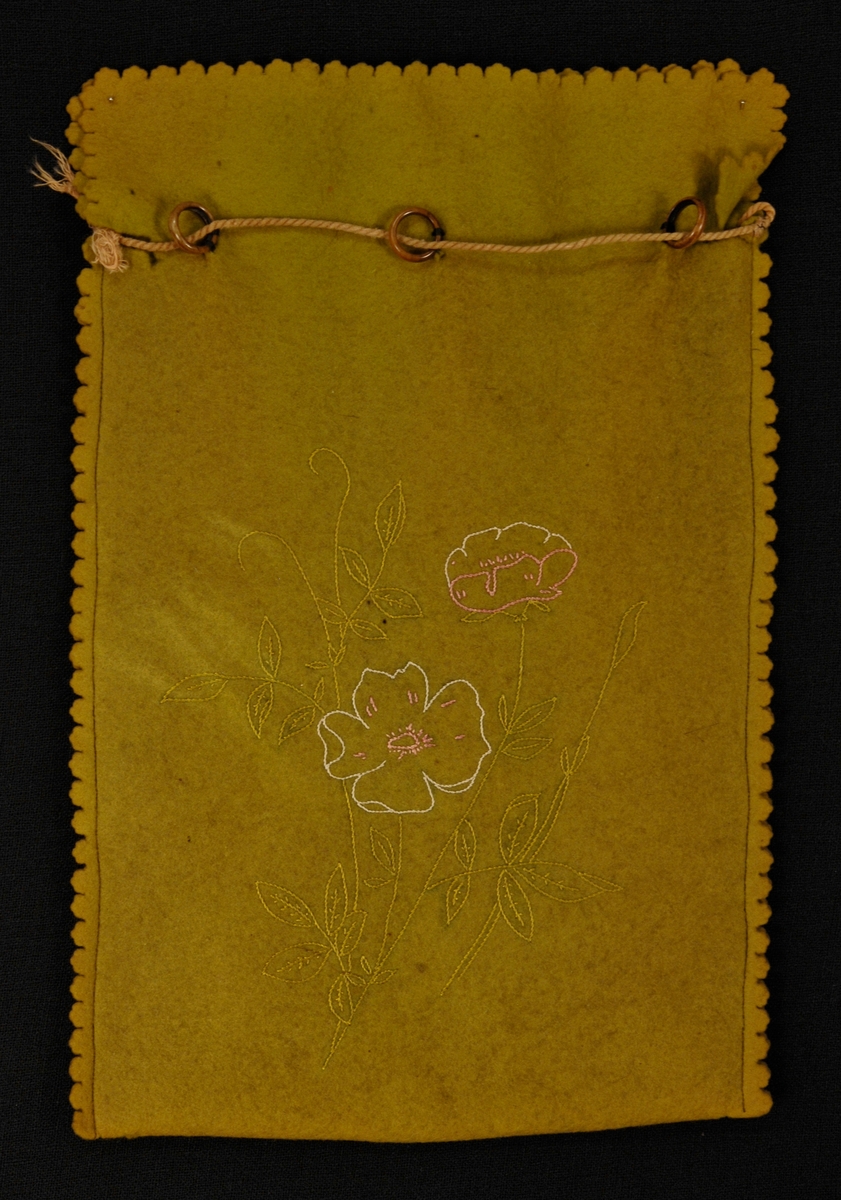 Pose i filt med broderi på eine sida, blader og to blomster, i øvre kant er det påsydd 6 beinringer. Filten er tagget langs sidene og oventil. Snor øverst.