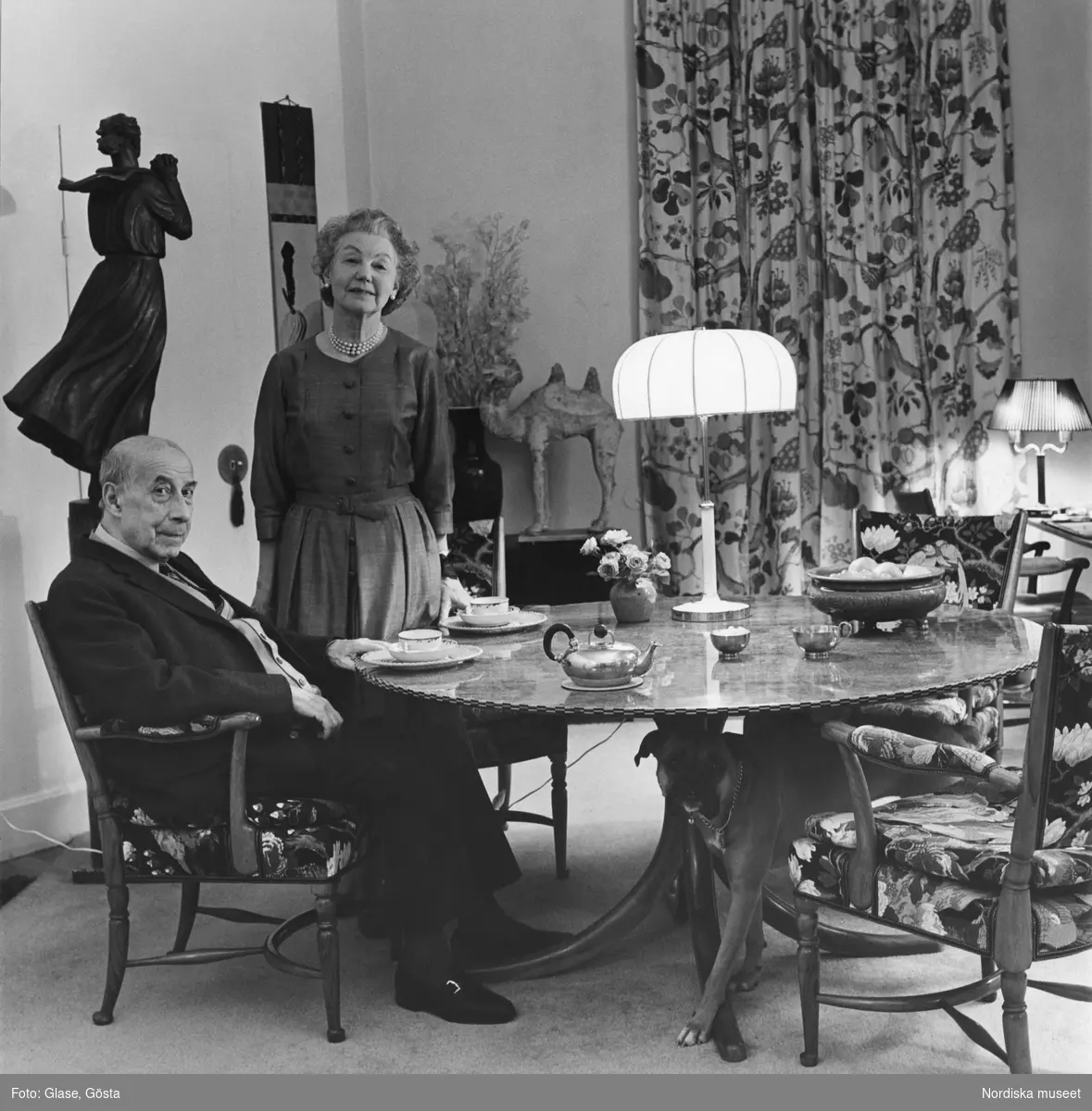 Svenskt Tenns grundare Estrid Ericson och hennes vän och medarbetare arkitekten Josef Frank tillsammans vid en matsalsmöbel. Bordet är dukat med tekoppar och kanna. Med på bilden är Estrids boxerhund.