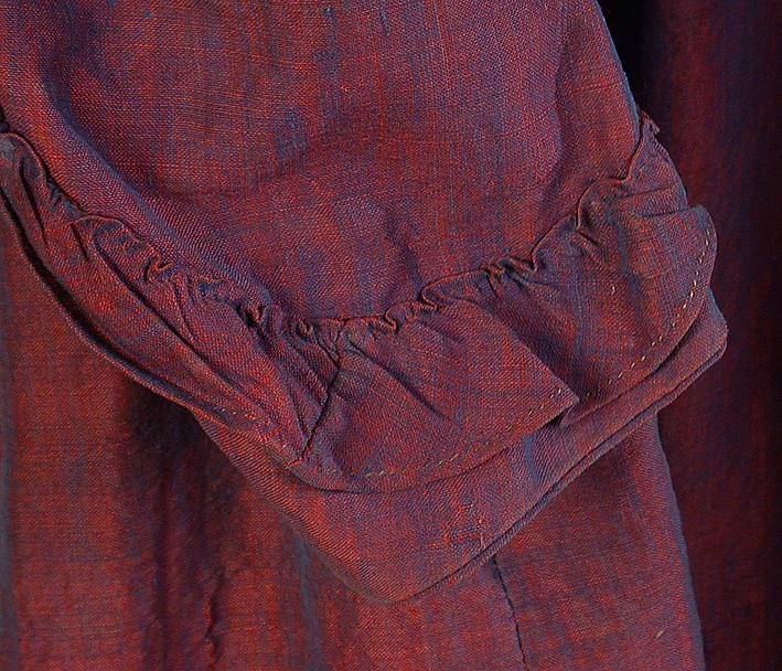 Rödlila klänning med skiftning av blått, av förr så kallad viktoriavävnad. 
Liv: Insvängt, insydda fiskbensfjädrar. Smal, rundad halslinning. Knäppning fram, med 7 st klädda träknappar. I brösthöjd på livets knäppning finns 5 st hyskor och hakar mellan knapparna. 2 st snedsydda insnitt för bättre passform på framstycket. Formsydd rygg i 3 st delar, dekorationssöm med svart tråd. Livet kantat nedtill. Lång tvåsömsärm isydd med passpoal. Påsydd, rynkad volang nedtill på ärmen. Hänghällor fastsydda på baksidan av båda ärmhål. Troligen har liv och kjol varit delade, men är nu hopsydda.
Mått: Rygg 40 cm, ärm 57 cm.

Kjol: 7 våder. Rynkparti mitt bak, lagda veck i sidorna, slät fram. Kantad med brickvävda mörkgröna ylleband nedtill. Sparkband av beige- och brunrandigt tuskaftat linnetyg med skarvdel av enfärgat linne.