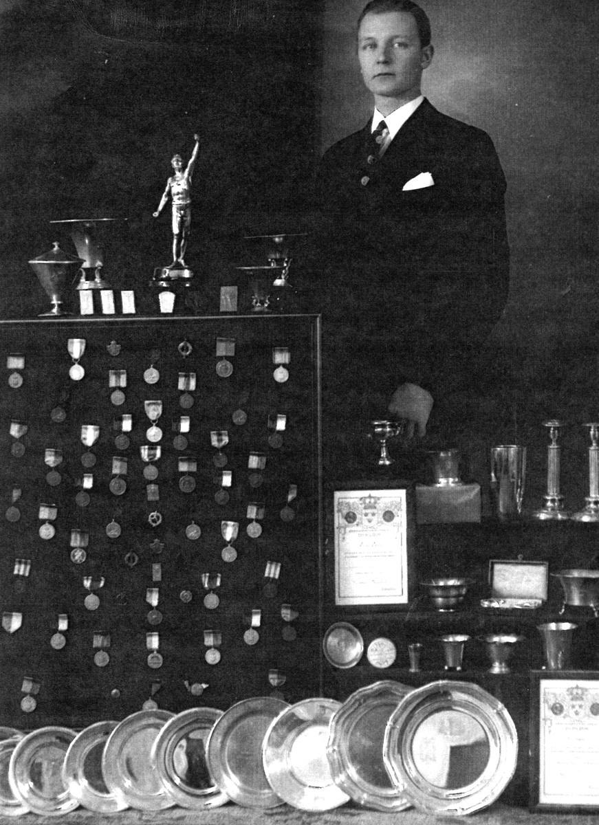 Nils Holm stående vid sin prissamling som aktiv inom KIS. Nils Holm var bl a spjutkastare, och medverkade tillsammans med tre andra Köpingsgymnaster vid OS i Berlin 1936.