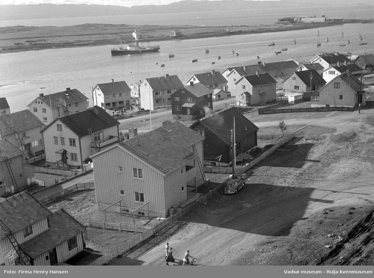 Vadsø 1957. Bildet er tatt fra kirken som sto ferdig i 1958. Bildet er tatt mot sydøst og viser boliger langs Damsveien, Nyborgveien og opp langs Tollbugata. Vi ser også litt av havna og Vadsøya. Hurtigruta er på vei inn i havna og vi ser fiskebåter og småbåter ankret opp i havna. På øya ser vi bygninger og kaier, fiskebruk. I høyre billedkant, mot vest ser vi Vadsø Sildoljefabrikk.
