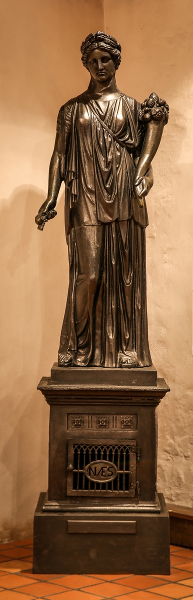 Statuen står på en firkantet sokkel, fyrkammeret, med firkantet gitterdør merket med NÆS. Statuen er en kopi av drapert antikk kvinnefigur, kronet med hvetekrans og overflødighetshorn i sin venstre og kornaks i sin høyre hånd. Avtrekk gjennom ryggen.