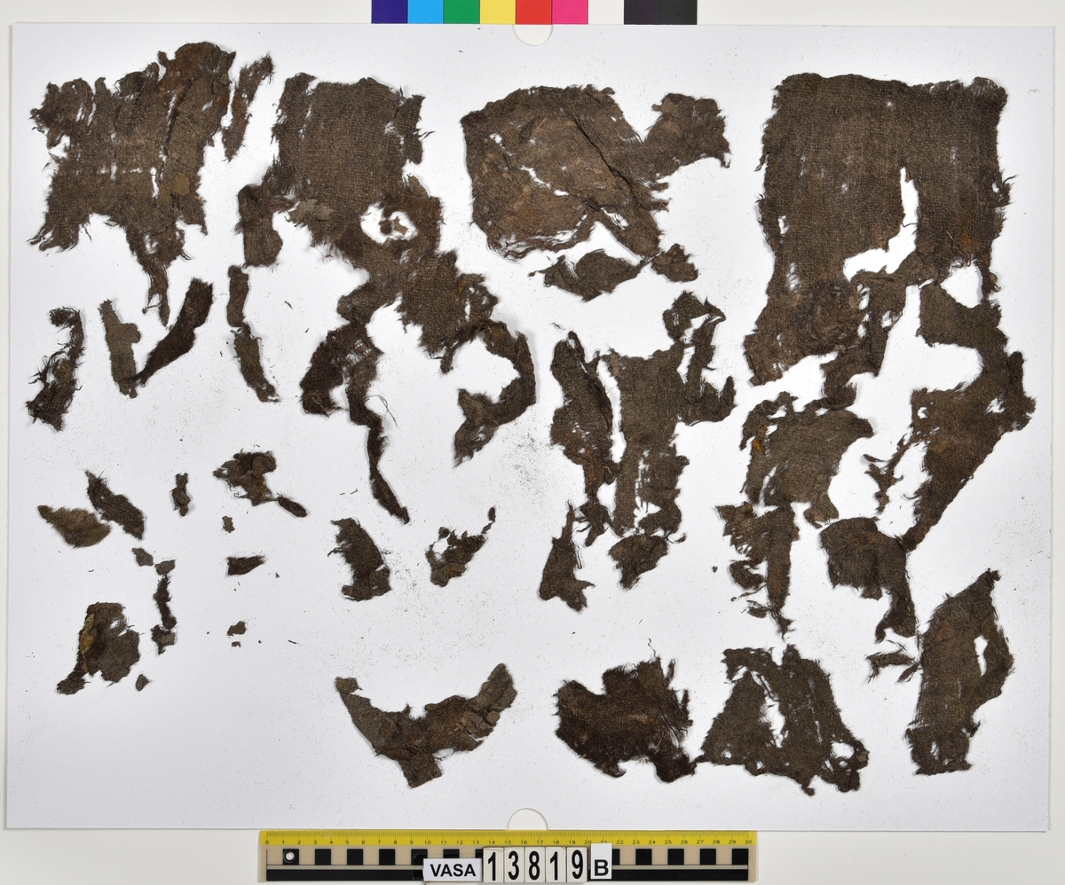 Textilfragment, byxa.
125 fragment uppdelade på fyndnummer 13819a-d.
Fnr 13819a består av 44 fragment av ull vävt i tuskaft och valkat på ena sidan. En del av fragmenten har originalkanter med sömmar och fållar.
Fnr 13819b består av 35 fragment av ull vävt i tuskaft. En del av fragmenten har originalkanter med sömmar.
Fnr 13819c består av 40 fragment av ull vävt i tuskaft. En del av fragmenten har originalkanter med sömmar. Fragmenten är mycket sköra.
Fnr 13819d består av 6 fragment mönstervävt band av ull.