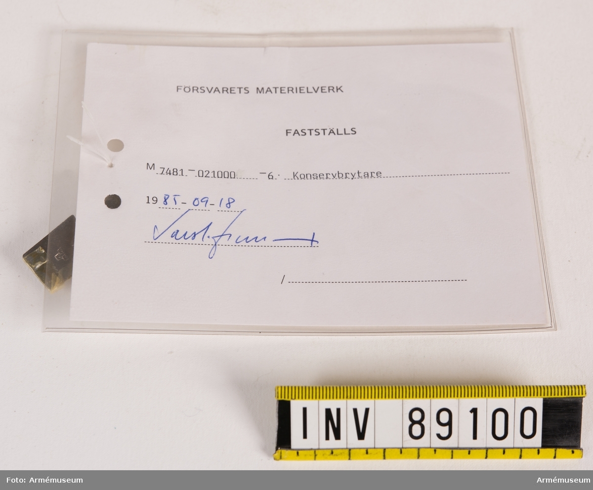 Konservbrytare i stål med tre kronor-stämpel. Vidhängande modellapp med text: "Försvarets materielverk. Fastställs. M 7481-021000-6 Konservbrytare. 1985-09-18. (oläslig underskrift)."