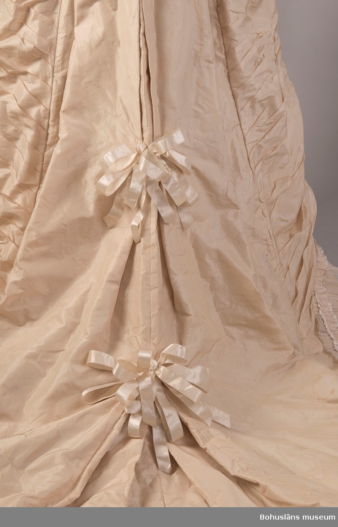 Gulvit klänning buren av Pauline Ekelund, född Berg (1843 - 1881), vid giftermål 1879 med överstelöjtnant Frithiof Ekelund. Fotografi från bröllopet, se bildarkivet UMFA samt ramat färglagt porträtt UM018507.
Inprovat liv, avskuren i sidorna, öppen fram, knäpps med tygkädda knappar. Halskant garnerad med gulvit, veckad bomullsspets, maskintillverkad samt kant av veckat siden med uppfransade kanter. Lång ärm avslutad med volang av tyget. För övrigt garnerad på samma sätt som kring halsen. Rosett av gulvitt sidenband fastsytt vid handleden. Lång kjol med släp. Rynkad mitt fram och vid sidorna. Baktill är kjolen skuren för turnyr. Släp garnerat med tre rosetter av gulvitt sidenband. Kjolkanten garnerad på ovansidan med veckat, tunt bomullstyg samt veckad remsa av tyget med uppfransade kanter. Under kjolfållen veckat bomullstyg som på ovansidan, kantad med smal, veckad kant, kantad med vit spets. Kjolfållen skodd med stärkt, vitt bomullstyg. Livet fodrat med vitt bomullstyg. 

Se bilagepärm med kopia av "inbjudan till vigselakten mellan Kaptenen vid Kongl. Bohus Läns Regemente Frithiof Ekelund och Pauline (född Berg) tisdagen den 7 oktober 1879 i Rådhussalen, Uddevalla". 

Ur punktnummerkatalogen 1957-1978:
Fru Anna Barks, sterbhus