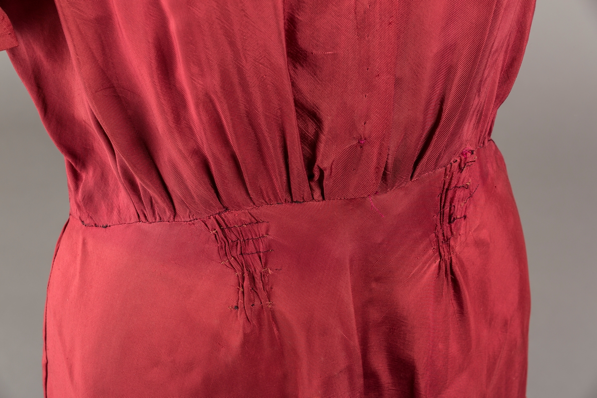 Form: Glatt, klokkeformet skjørt. Forstykkene rynket til buede bærestykker foran. Overdel rynket til skjørt. Korte ermer. Lukket med trykknapper. Belte fastsydd.