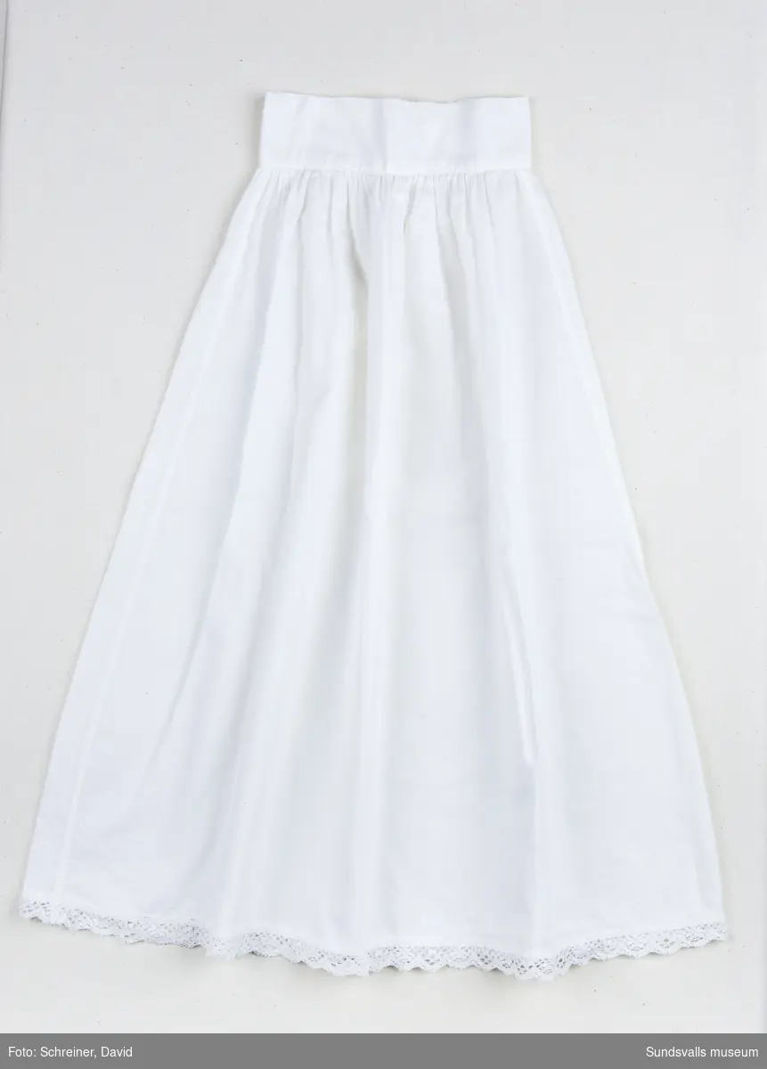 En vit bomullskjol med bred linning som knäpps baktill med två vita knappar. Kjolens nederkant har dekorerats med en brodyrspets.