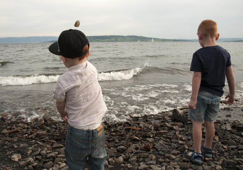 Two young boys dressed in shorts and T-shirts, one with a cap on his head, stand on a pebbly beach facing towards the water and throwing stones into it.