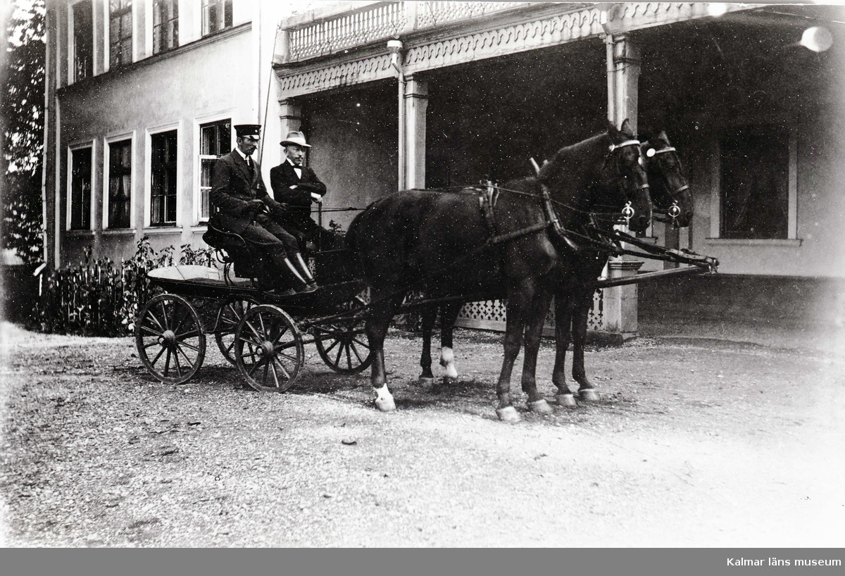 Framför huvudbyggnaden står en liten vagn dragen av parhästar. På kuskbocken sitter en kusk i uniform och en herre.