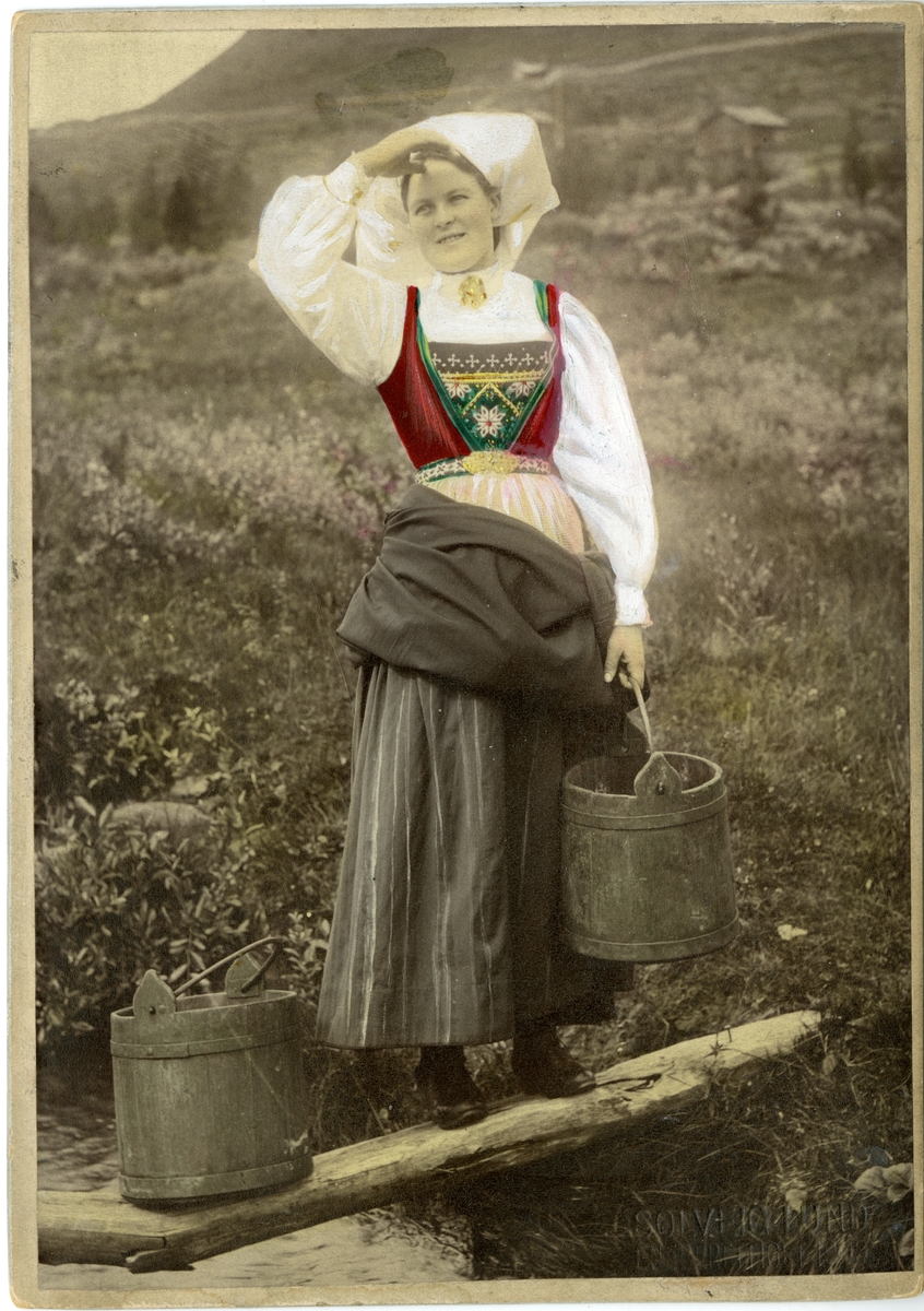 Kolorert fotografi av kvinne med drakt, på stokk over bekk bærende på to bøtter. Hardanger.