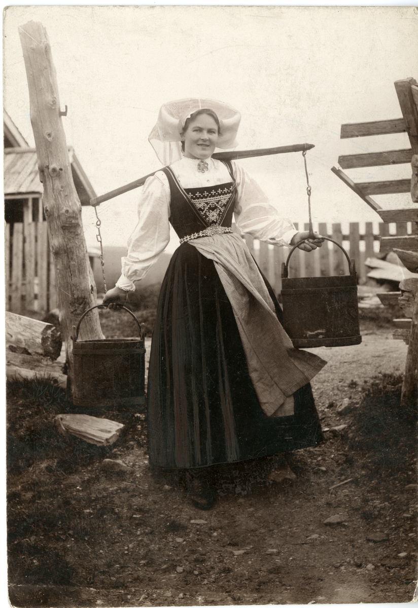 Fotografi av kvinne med drakt, på tun med åk og to vannbøtter. Hardanger.