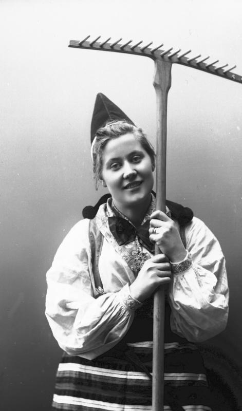 Kvinne i drakt og lue, som holder rive. 1908. (Foto/Photo)