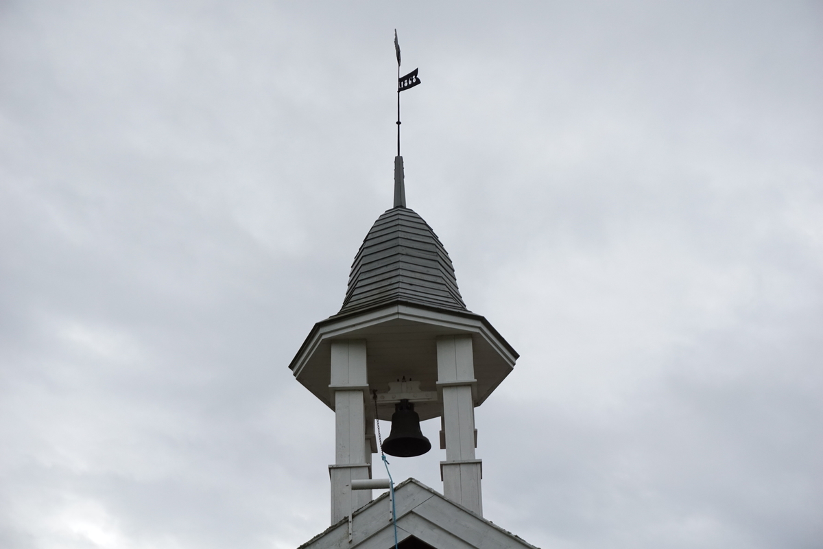 Helgeby, Nes, Ringsaker. Klokketårnet står plassert på gårdens stabbur, og har etter hva eier vet alltid stått på samme sted. Tårnet har en hvitmalt grunnkonstruksjon og et åttekantet hjelmformet tak kledd i spon.