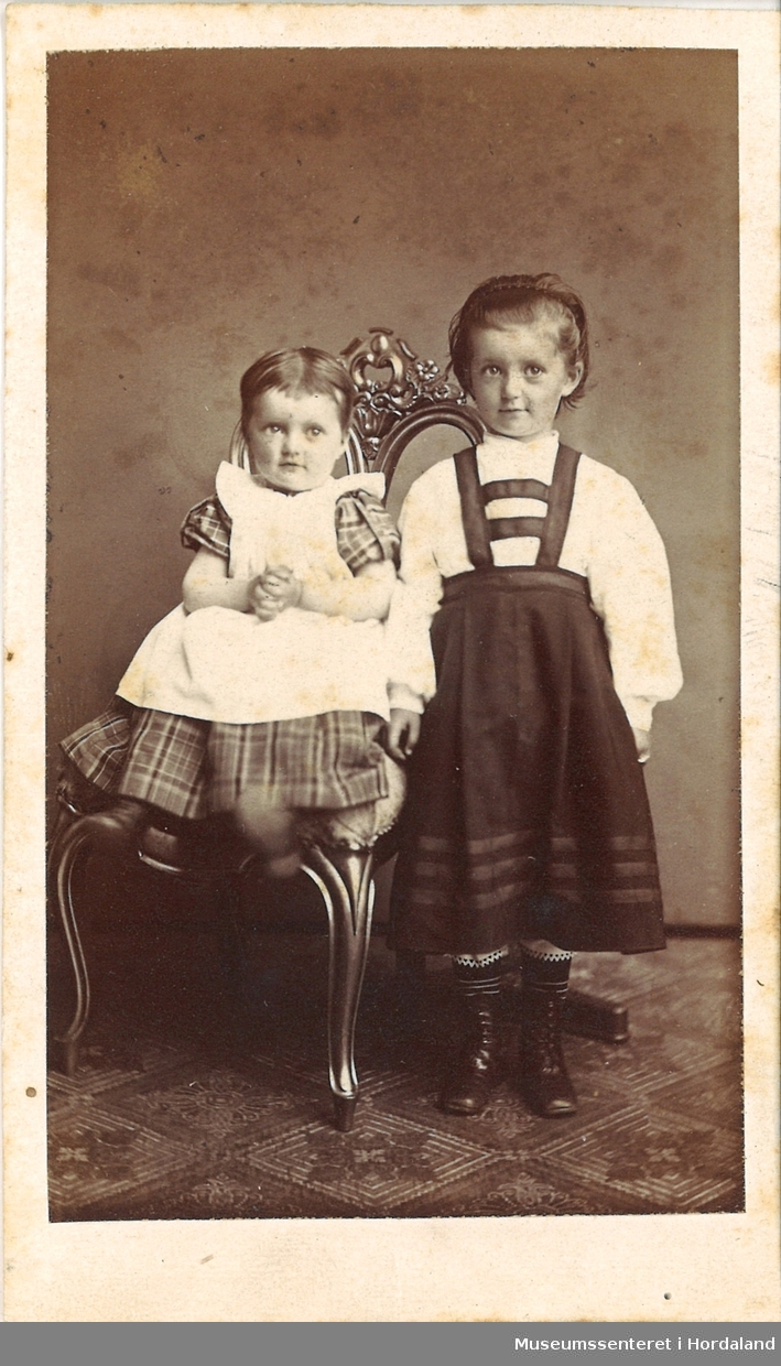 atelierfotografi av to små jenter, ei som sit på ein stol kledd i ruta kjole og forkle, ei litt større jente står til høgre med kvit bluse og seleskjørt