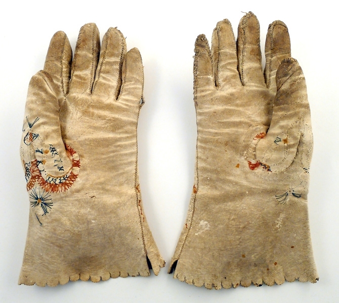 Handskar av vitt skinn med broderat mönster i grönt och två nyanser brunt. Även brun rand runt fingersömmarna. Broderiet delvis bortslitet. Smutsiga
