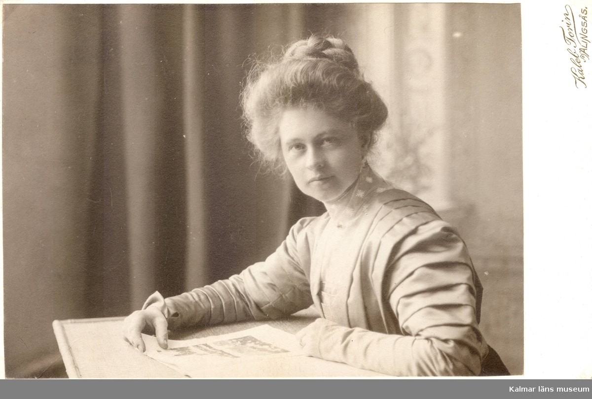 Ellen Warholm med pösigt, uppgummat hår och klänning med många veck, sittande vid ett bord. "Mösseberg 1908".