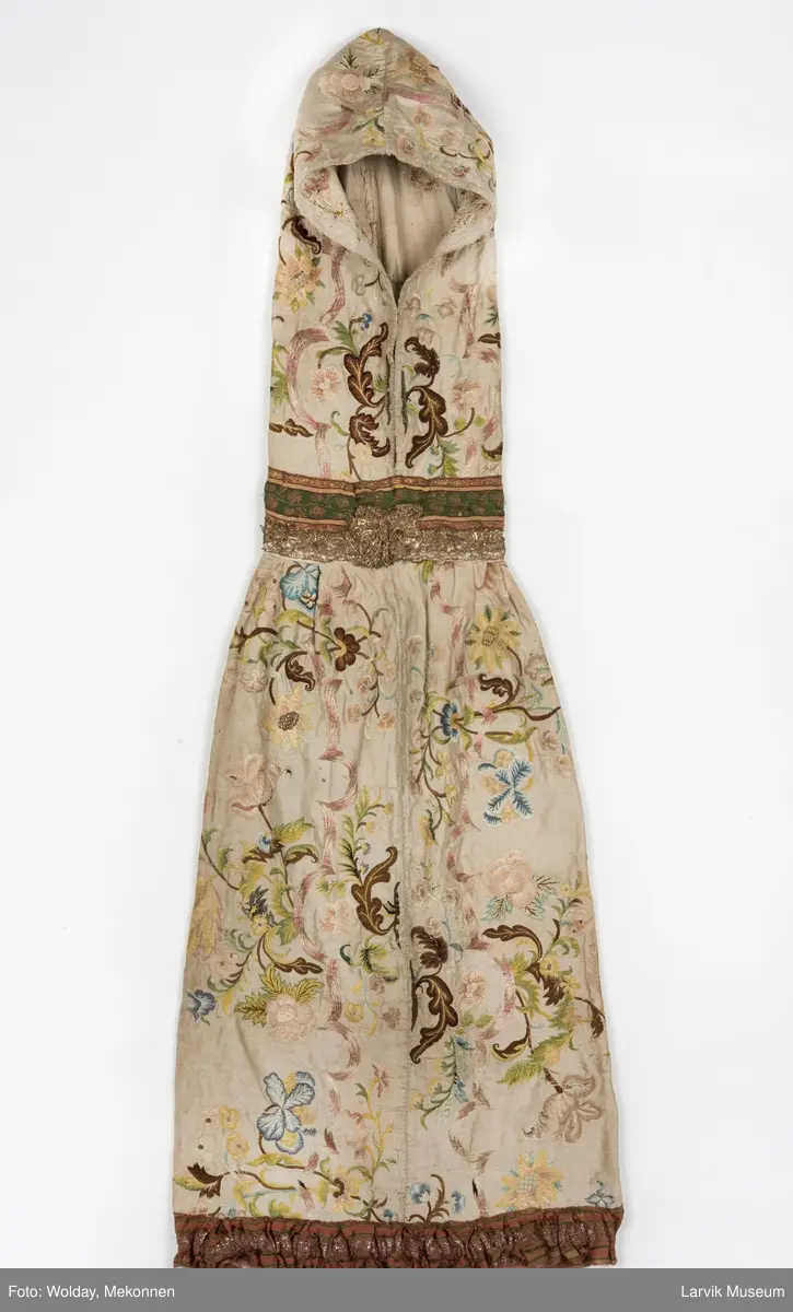 Form: livstykke som hette, åpen til livet, bred linning, rynket skjørt med blondekant nederst, søm bak på hetten i linning, søm foran og bak i skjørt, enkelte skjøtesømmer, linning brodert med gulltråd, skjørtet er foret med tynt stoff