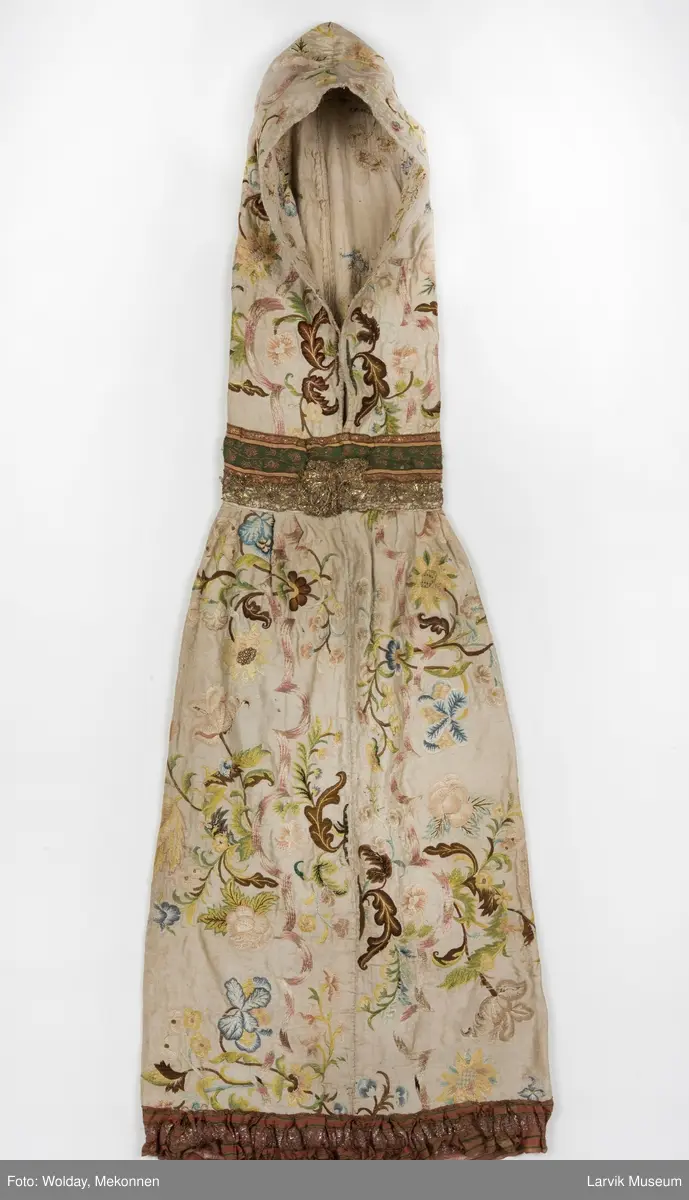 Form: livstykke som hette, åpen til livet, bred linning, rynket skjørt med blondekant nederst, søm bak på hetten i linning, søm foran og bak i skjørt, enkelte skjøtesømmer, linning brodert med gulltråd, skjørtet er foret med tynt stoff