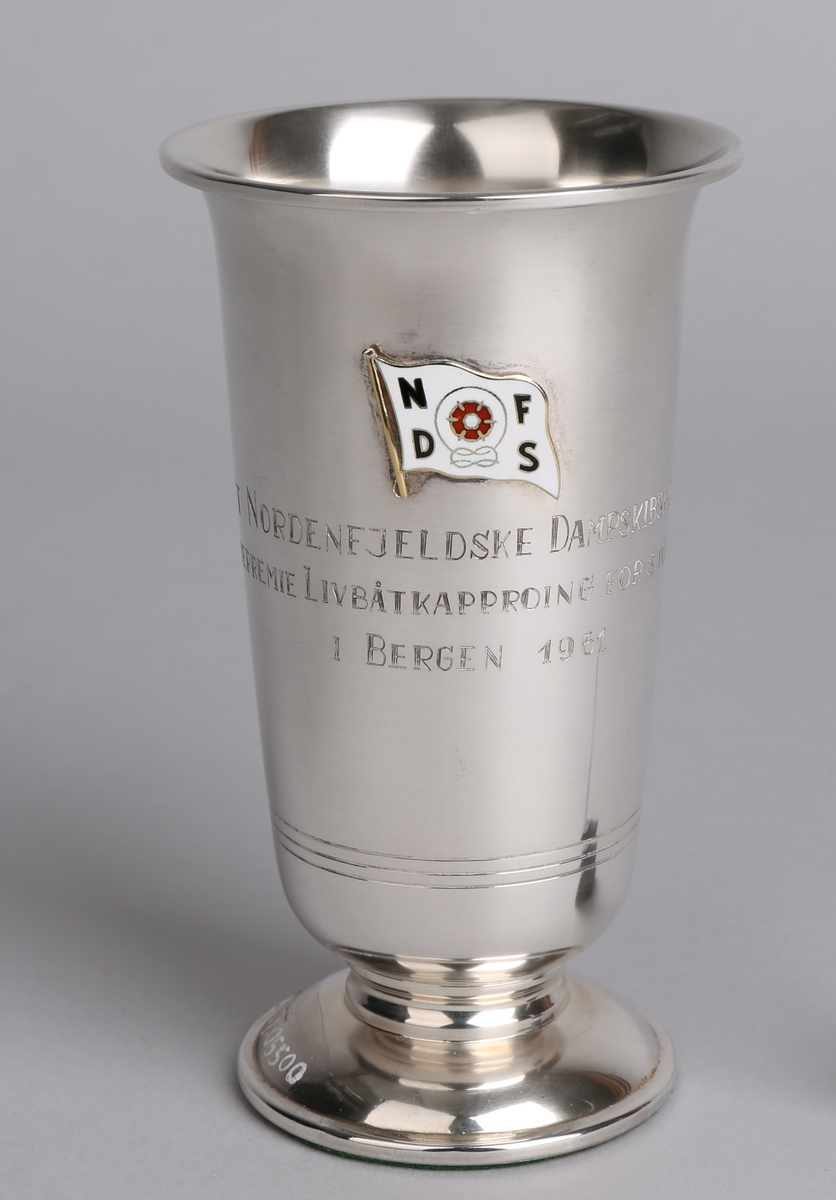 Sølvpokal fra Det Nordenfjeldske Dampskibsselskab, på sokkel med tekst inngravert samt emaljert rederimerke.
