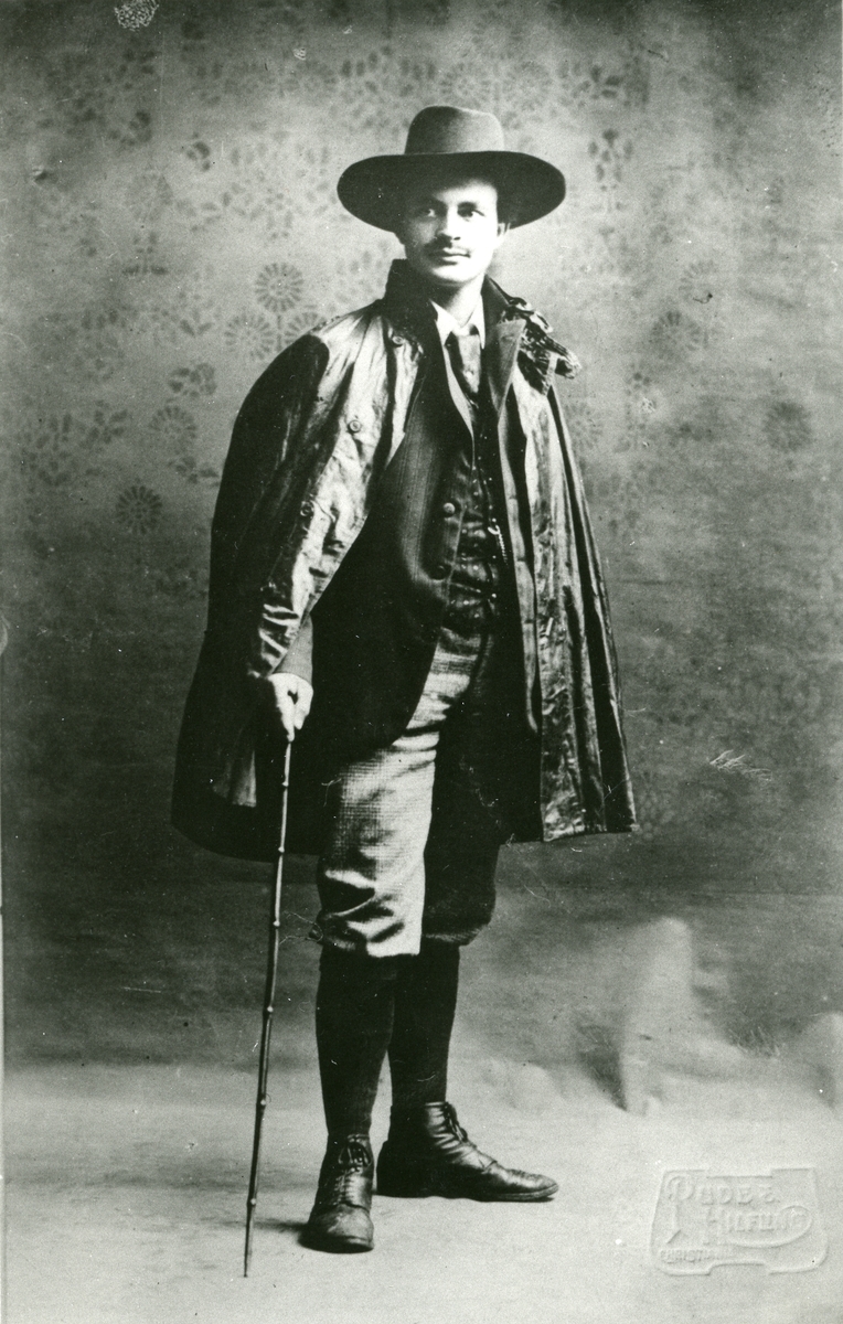 Mann oppstilt for fotografen m/hatt, spaserstokk og regnkappe over skulderen. Portrett av Haakon Garaasen (6/7 1887 - 1957)