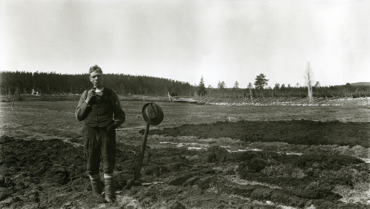 Mann, Otto Granli (1896 - 1930), stående i åker i Kallberg (Grønberget) med pipe i munnen. Hatten hans henger på hakka (el. lign.) som står i jorda ved siden av ham.
