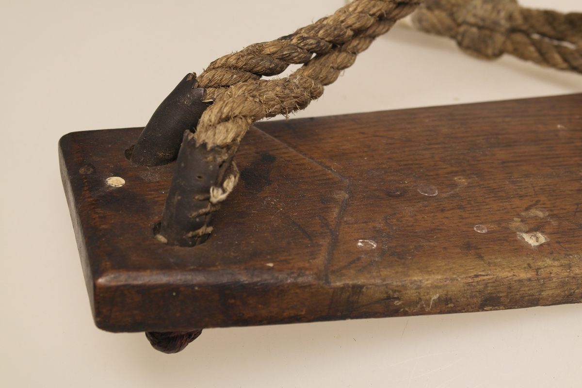 Båtmannsstol i skåret, høvlet og pusset eik. Rektangulært brett som smalner i endene. Brettet er buet. Oppheng består av to tau i tvunnet bast. Tauene er festet i hvert hjørne av brettet. De er beslått med lær for å beskytte mot slitasje mot treet. Læret er sydd i sammenføyningene. Tauene er samlet rundt et sinkbeslått "øye". Det er surret bomull rundt tauene som går rundt "øyet".