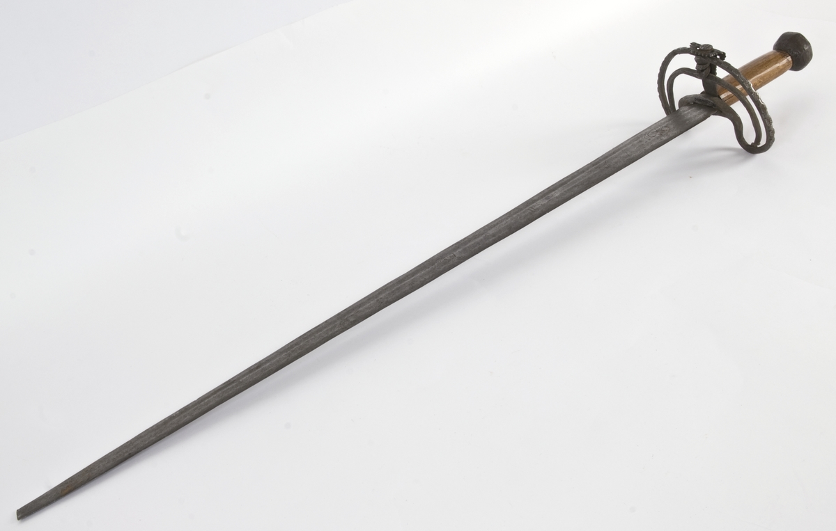 Fasettert rundt knapp med 8 fasetter. Det er innslått 4 smedmerker på knappen, i annethvert felt. Tregrep  Asymerisk åpen parerring med  bøyler med rektangulært tverrsnitt og med kringlefasong med en ytre og en indre parerring. Ytre ring har utsmidde rektangulære partier med kryssfliling. Ytre og indre ring er låst ved at de er tvunnet sammen i senter. Utsiden er smidd som en plateformet deksel. Denne typen pareranordning er trolig norsk
Bred tveegget klinge med 3 brodrender på hver side. Klingen er merket med  Solingens løpende ulv, et hjul og 2 andre symboler på begge sider. Klingen er brukket.