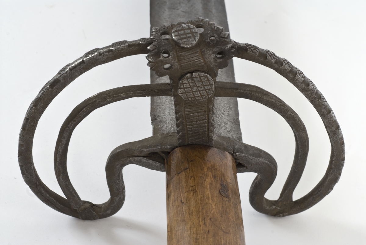 Fasettert rundt knapp med 8 fasetter. Det er innslått 4 smedmerker på knappen, i annethvert felt. Tregrep  Asymerisk åpen parerring med  bøyler med rektangulært tverrsnitt og med kringlefasong med en ytre og en indre parerring. Ytre ring har utsmidde rektangulære partier med kryssfliling. Ytre og indre ring er låst ved at de er tvunnet sammen i senter. Utsiden er smidd som en plateformet deksel. Denne typen pareranordning er trolig norsk
Bred tveegget klinge med 3 brodrender på hver side. Klingen er merket med  Solingens løpende ulv, et hjul og 2 andre symboler på begge sider. Klingen er brukket.