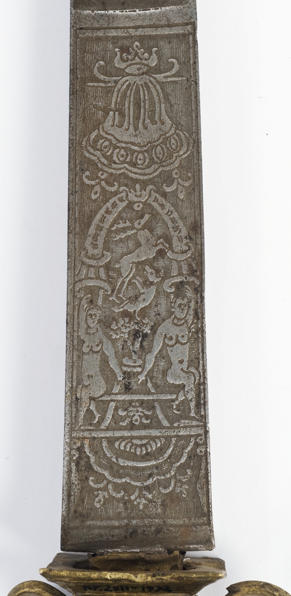 Messing feste fra en kårde. Rund fasettert knapp med nitteknapp.
Tregrep med snodd kobbertråde bevikling og bånd oppe og nede. Fremre parerstang brukket Bakre parerstang går over i håndbøylen, som er tappet inn i knappen.Parerplaten er fjernet, rektangulært baisstykke.
Rett enegget klinge med kileformet tverrsnitt og to smale ryggstillt blodrender.
Klingen har etset dekor med nanvgitte figurer og andre jaktscener.