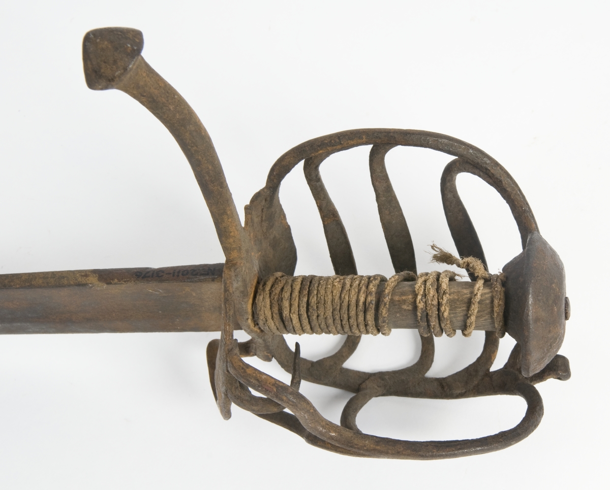 Soppformet  knapp. Tregrep med trådbevikling.  Runde parerstenger og svakt nedbøyd bakre parestang. Fremre parerstang er brukket. Kurvfeste med runde bøyler med utsmidde  midtpartier. Nyreformet parerplate med tombøyle og klingebrekker på innsiden. 
2 sidebøyler på innsiden.
Enegget, kileformet krum klinge med  bred blodrand. Gjeddenebb og ryggslipning mot spissen.  
Klingens innside merket med 3 smedstempler.