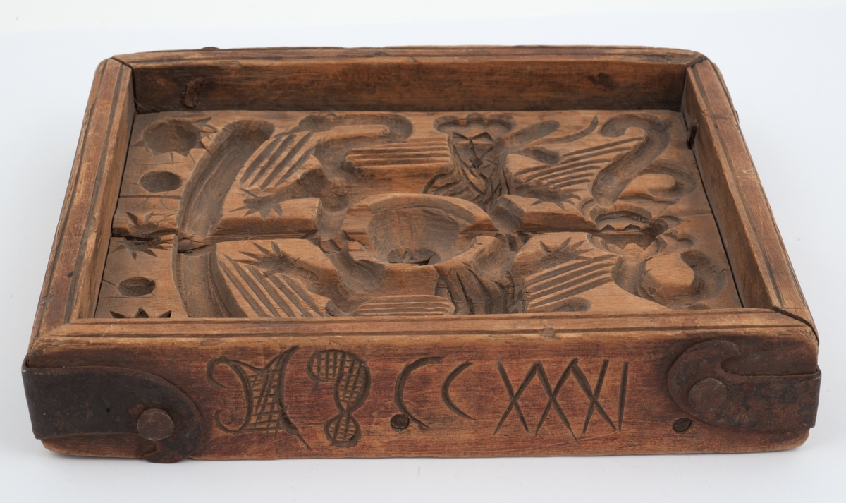 Firkantet kakeform med jernbeslag på hjørnene. Den ene siden av bunnen skåret med sol, måne, fabeldyr og et tre. Undersiden har blomster og rankedekor med 2 fugler. Litt skåret dekor på den andre siden.
Innskrift på den ene langsiden.