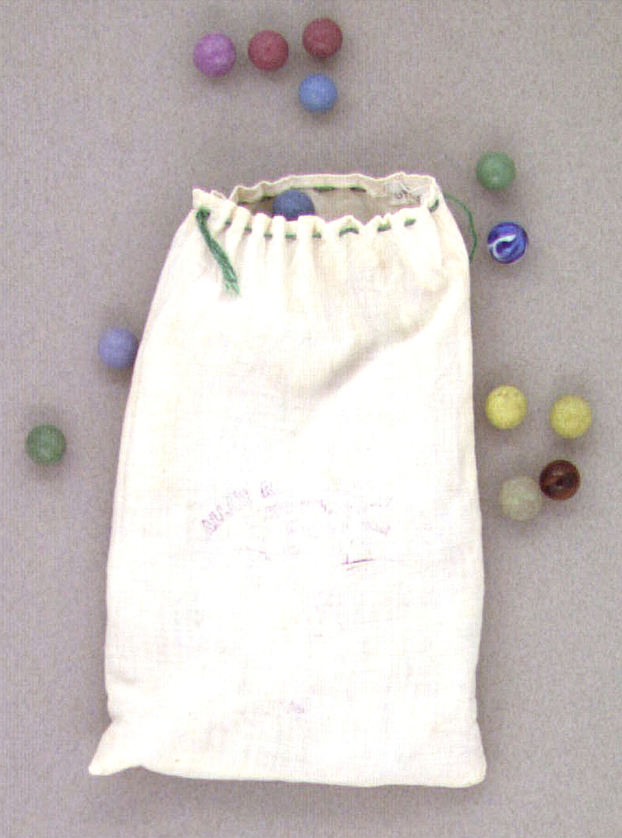 En vit tygpåse av bomullstyg innehållande spelkulor av stenmassa, kullager och glas. Påsen har oläslig text på ena sidan samt försedd med dragsko med grönt bomullsgarn.