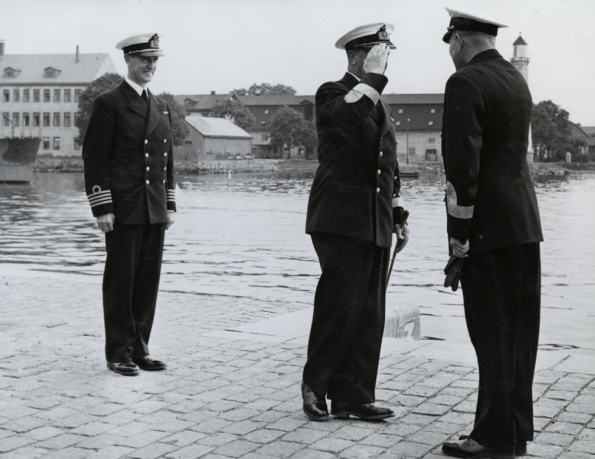 Konteramiral Samuelson gör en hälsning till konteramiral Berthelsson på Kungsbron. Bakom honom står kommendörkapten av första graden Bengt Lind af Hageby. Samtliga är iförda uniform. I bakgrunden syns Stumholmen och vattnet däremellan.