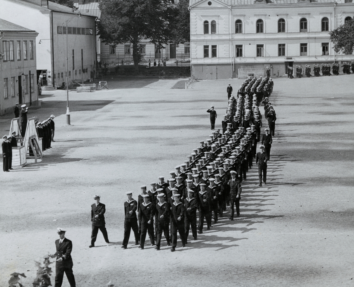 Uniformsklädda flottister marscherar över Sparre kaserngård under ledning av en kapten. I bakgrunden syns bataljon Sparres kanslibyggnad uppförd omkring 1818.